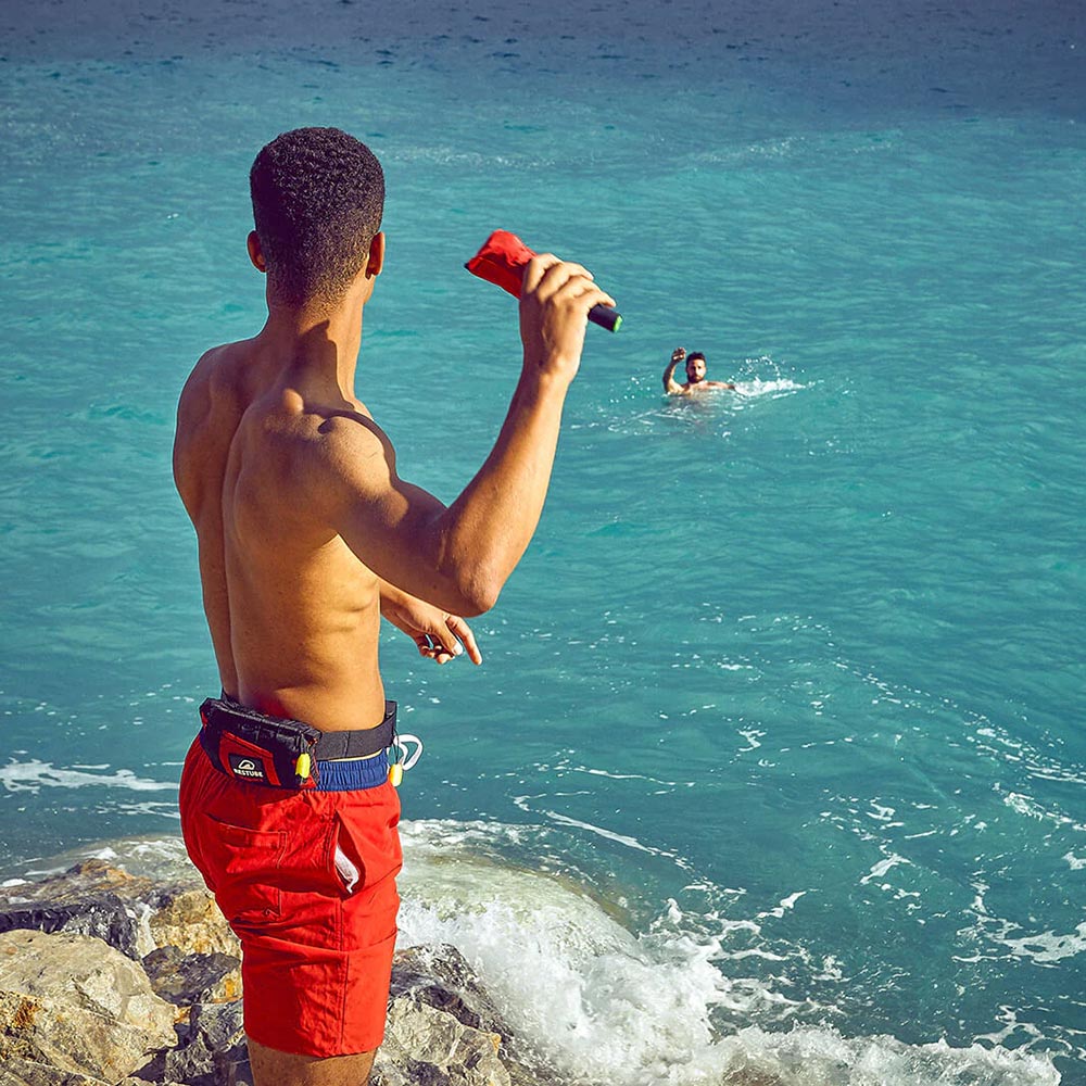 A man standing on a rocky shore ready to throw the Restube Automatic Water Rescue Buoy into the water to help a person needing assistance.